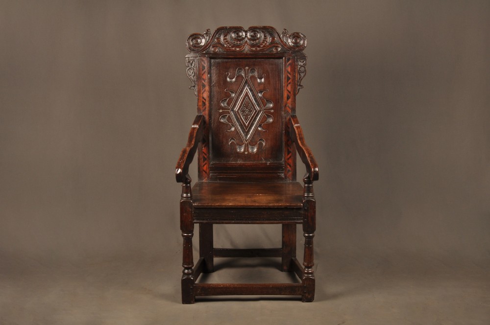 A seventeenth-century carved dark oak wainscot armchair featuring a carved lozenge motif on the back panel, scrolled cresting rail, shaped armrests, and turned front legs.