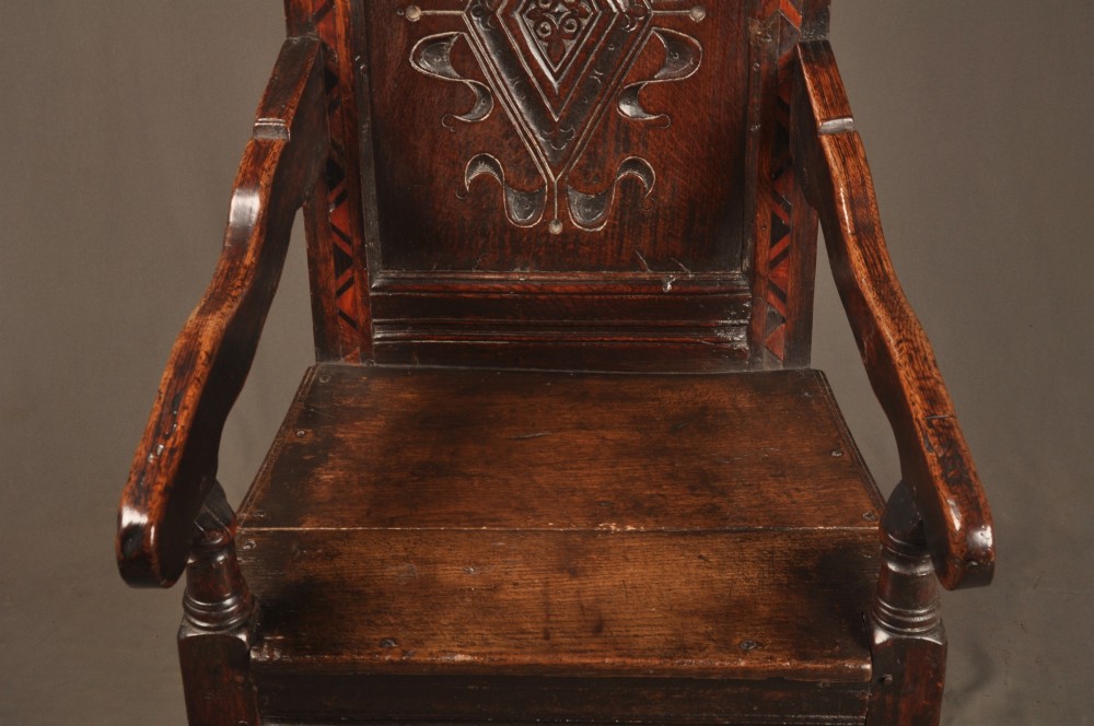 A seventeenth-century carved dark oak wainscot armchair featuring a carved lozenge motif on the back panel, scrolled cresting rail, shaped armrests, and turned front legs. View V