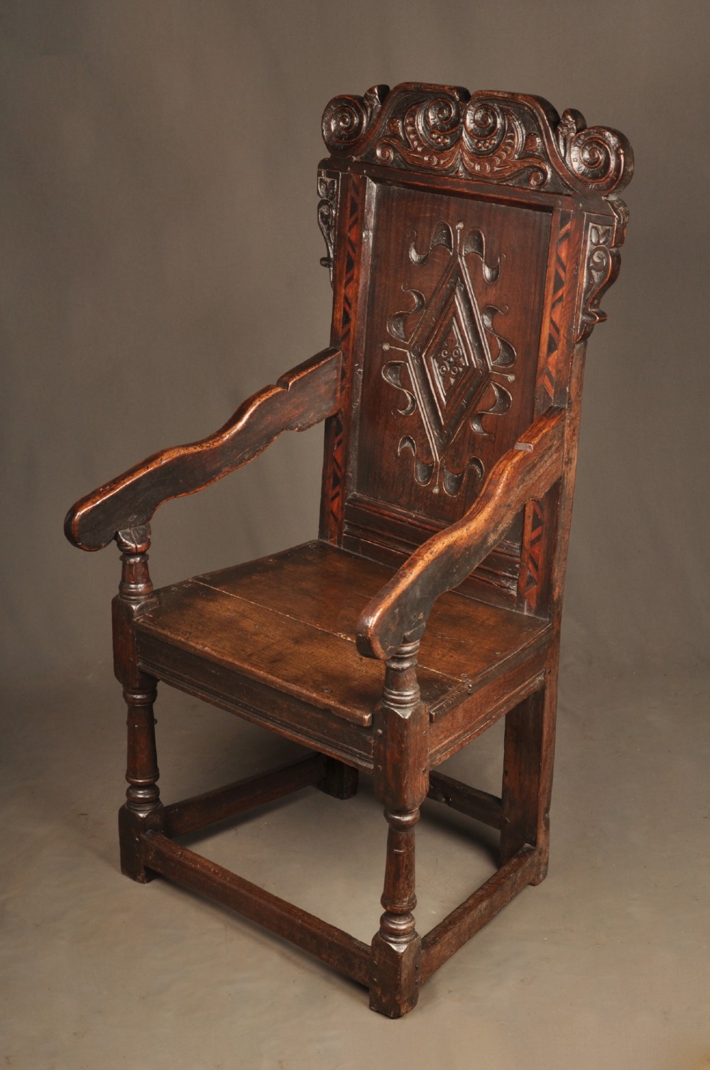 A seventeenth-century carved dark oak wainscot armchair featuring a carved lozenge motif on the back panel, scrolled cresting rail, shaped armrests, and turned front legs. View IX