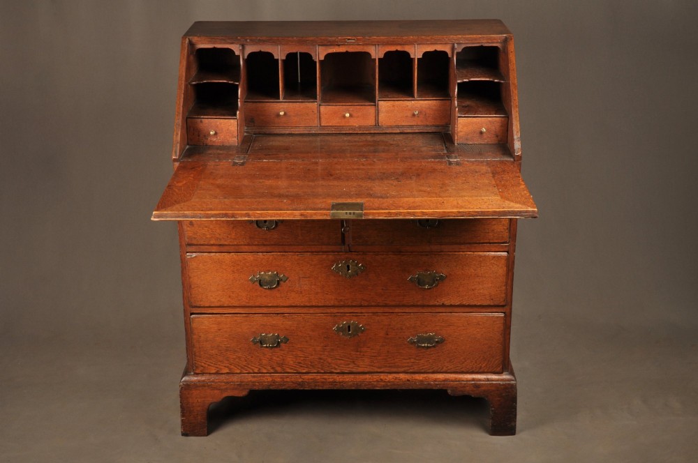 An early 18th-century oak bureau featuring a slant-front writing flap above two short and two long drawers with brass hardware. View IX