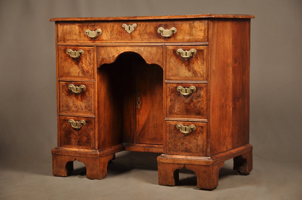 A Queen Anne walnut kneehole desk featuring figured wood veneers, seven drawers with brass handles, a central recessed cupboard, and bracket feet.
