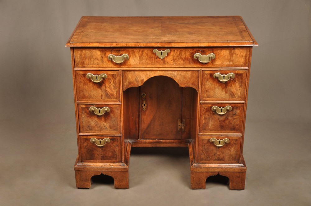 A Queen Anne walnut kneehole desk featuring figured wood veneers, seven drawers with brass handles, a central recessed cupboard, and bracket feet. View III