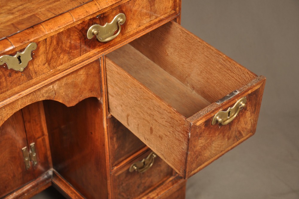 A Queen Anne walnut kneehole desk featuring figured wood veneers, seven drawers with brass handles, a central recessed cupboard, and bracket feet. View V