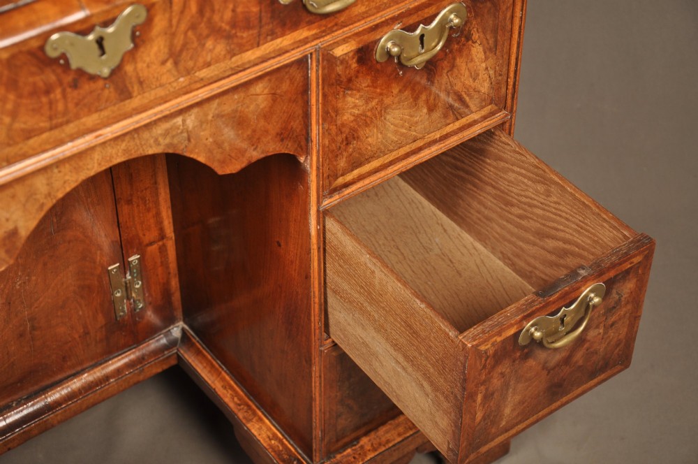 A Queen Anne walnut kneehole desk featuring figured wood veneers, seven drawers with brass handles, a central recessed cupboard, and bracket feet. View VI