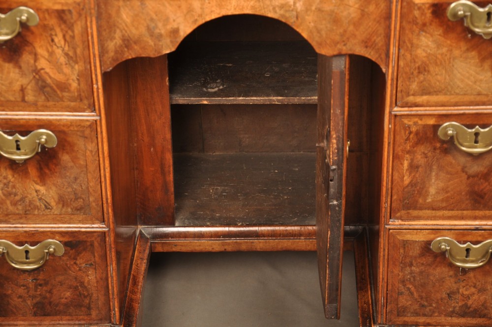 A Queen Anne walnut kneehole desk featuring figured wood veneers, seven drawers with brass handles, a central recessed cupboard, and bracket feet. View IX