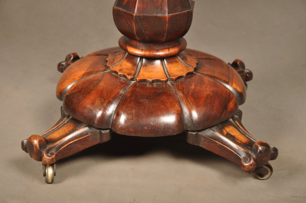 A Regency period rosewood fold-over card table featuring a carved apron, a central pedestal column, and a fluted quadripartite base on brass castors. View IV