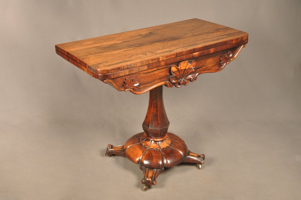 A Regency period rosewood fold-over card table featuring a carved apron, a central pedestal column, and a fluted quadripartite base on brass castors. View V