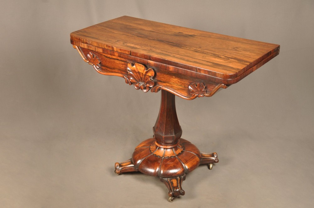 A Regency period rosewood fold-over card table featuring a carved apron, a central pedestal column, and a fluted quadripartite base on brass castors. View VI