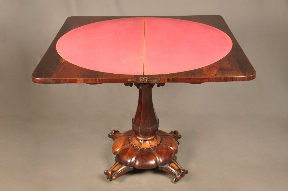 A Regency period rosewood fold-over card table featuring a carved apron, a central pedestal column, and a fluted quadripartite base on brass castors. View IX