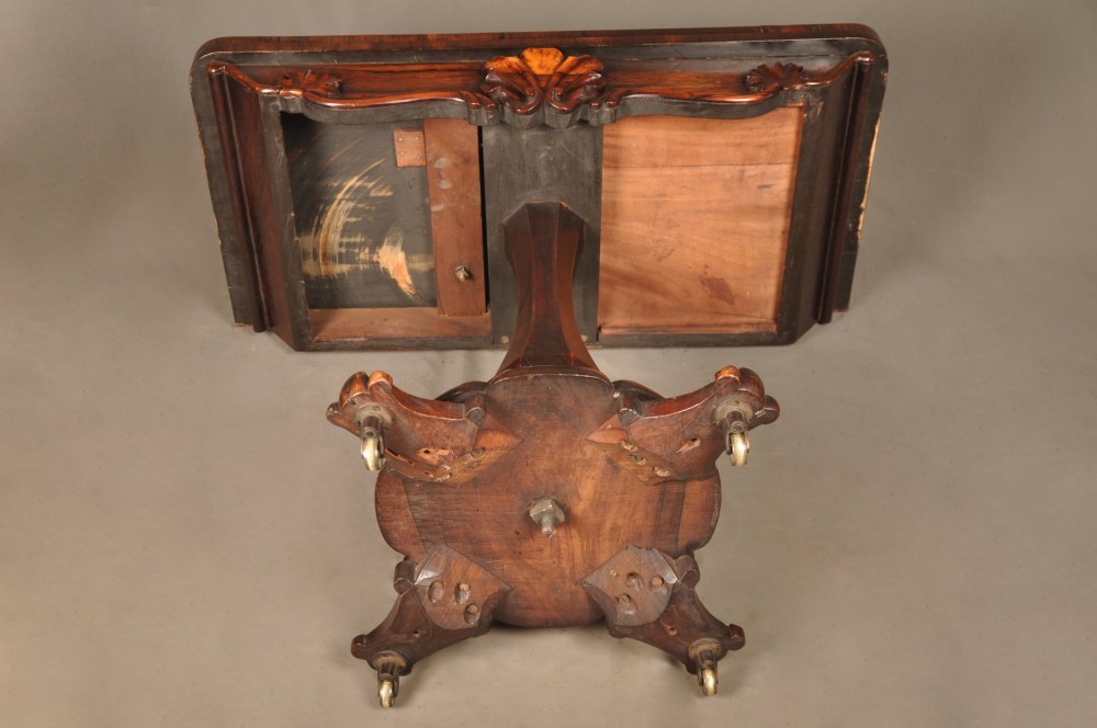 A Regency period rosewood fold-over card table featuring a carved apron, a central pedestal column, and a fluted quadripartite base on brass castors. View X