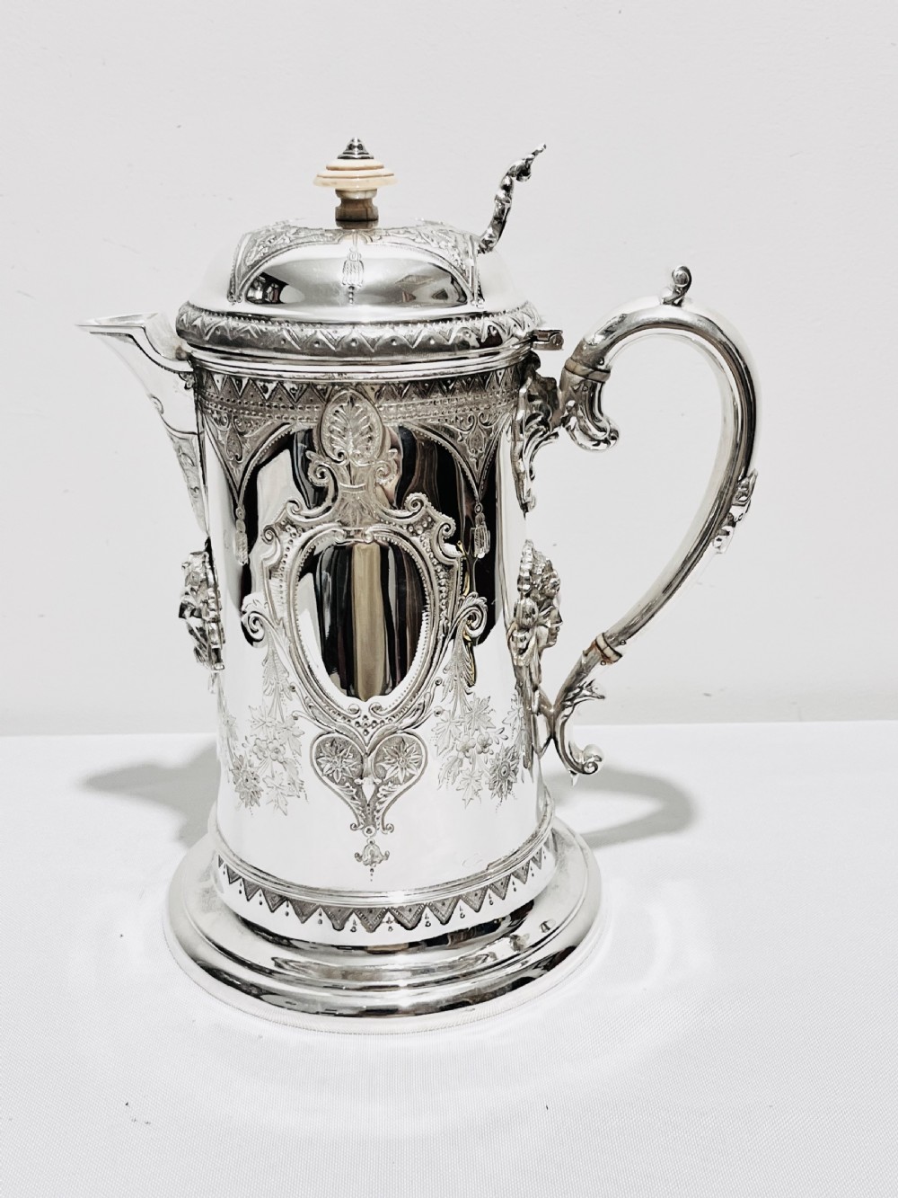 A Victorian lidded silver-plated jug featuring chased floral engraving, a scroll handle with insulating rings, and a hinged lid. View V