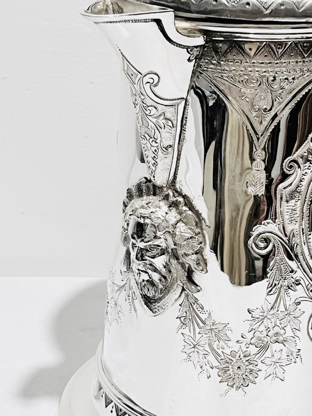 A Victorian lidded silver-plated jug featuring chased floral engraving, a scroll handle with insulating rings, and a hinged lid. View VII