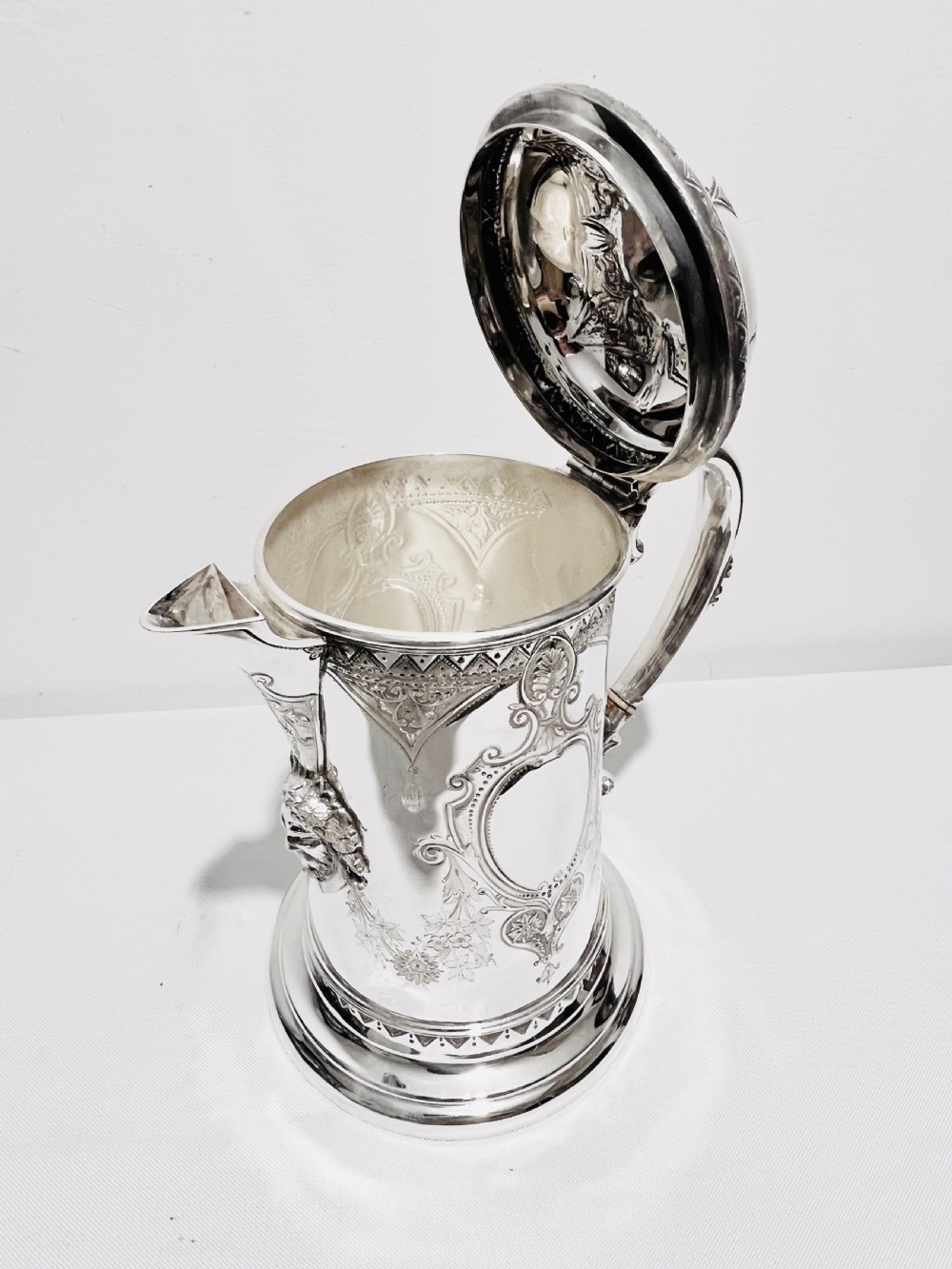 A Victorian lidded silver-plated jug featuring chased floral engraving, a scroll handle with insulating rings, and a hinged lid. View IX