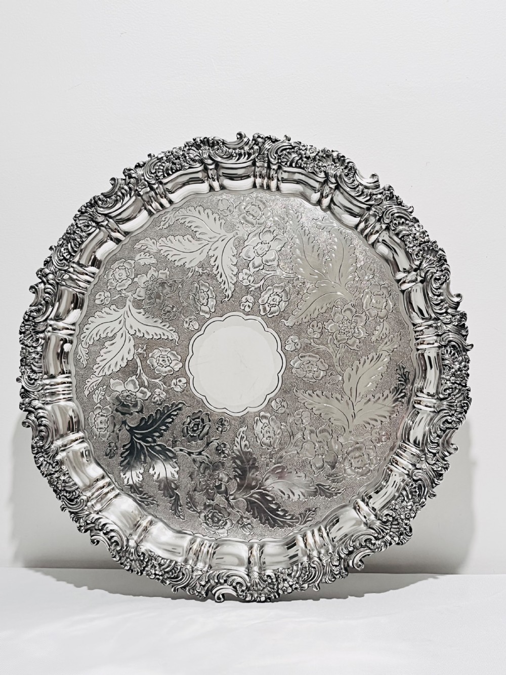 A circular silver plated salver featuring an ornate chased floral surface and a raised shell and scroll rim.