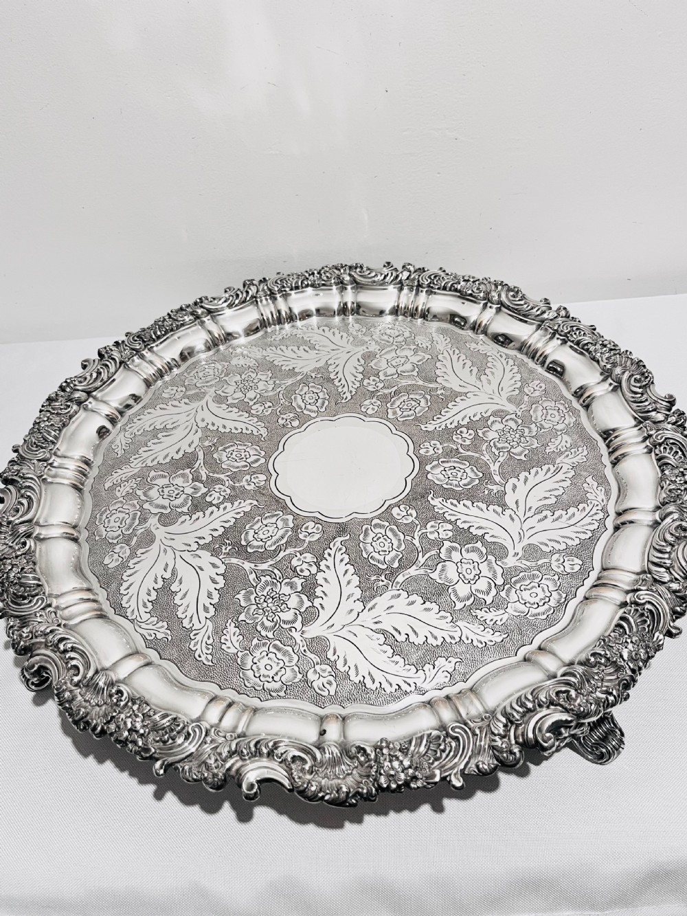 A circular silver plated salver featuring an ornate chased floral surface and a raised shell and scroll rim. View II