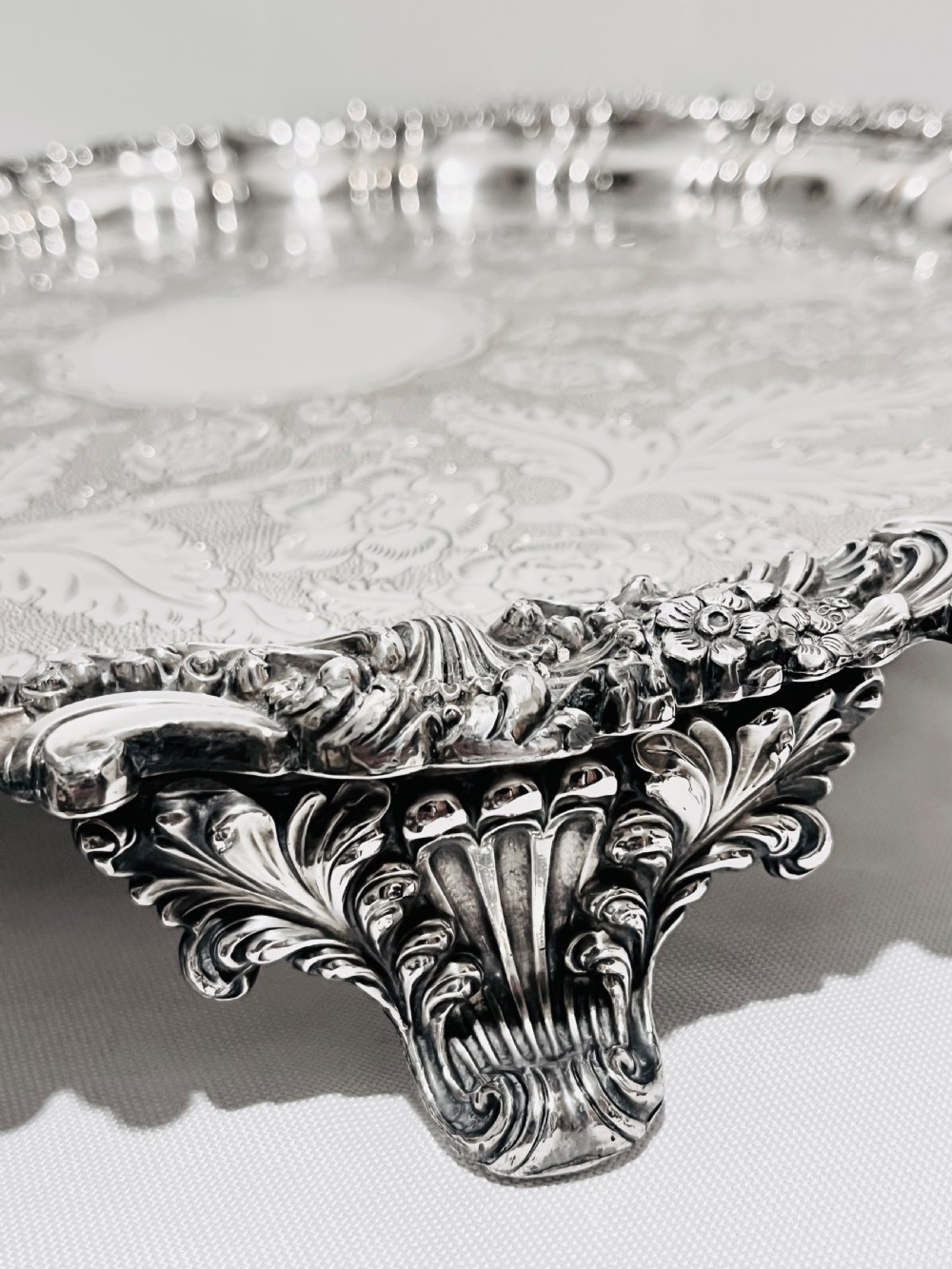 A circular silver plated salver featuring an ornate chased floral surface and a raised shell and scroll rim. View III