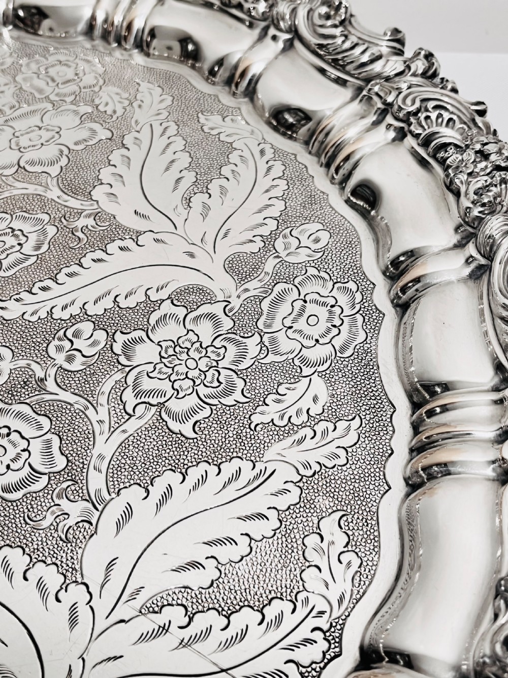 A circular silver plated salver featuring an ornate chased floral surface and a raised shell and scroll rim. View V