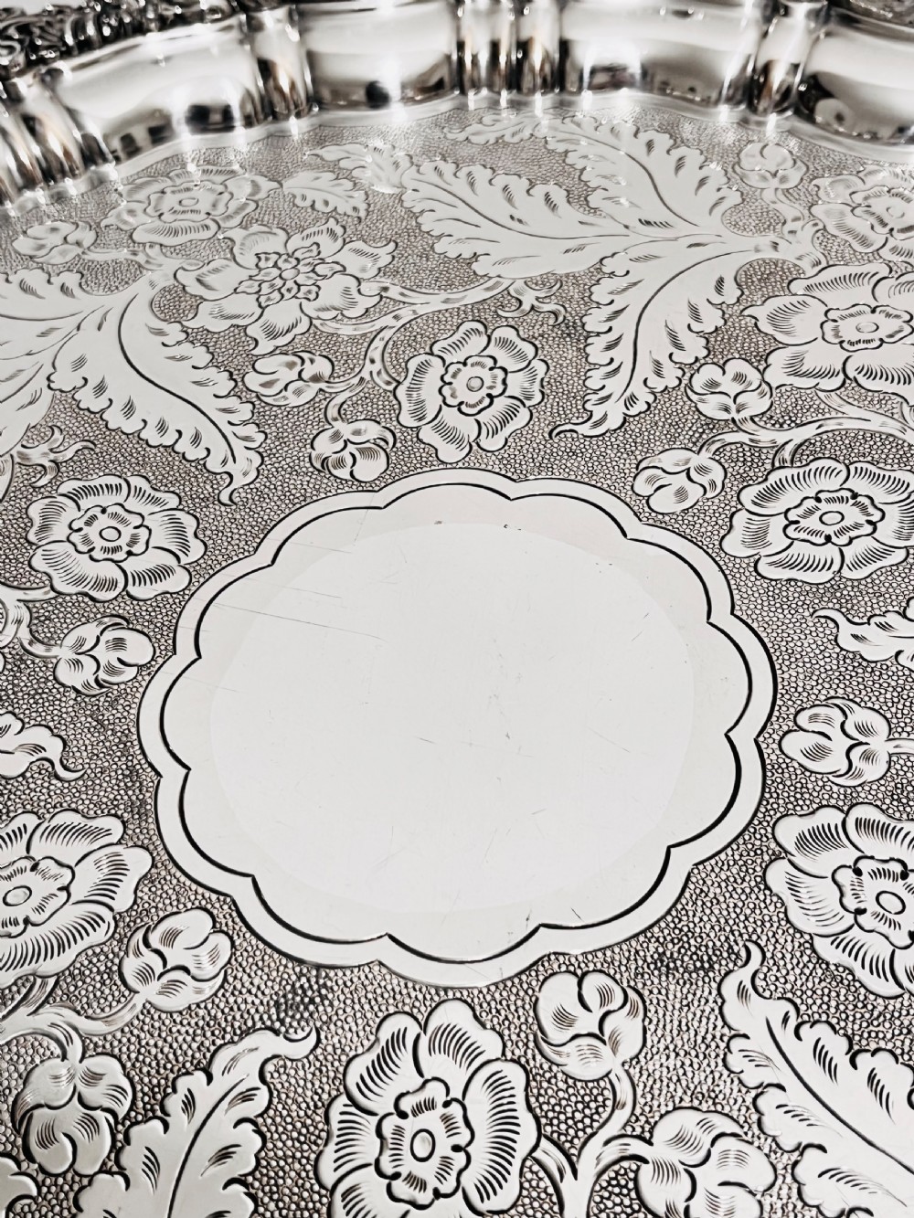 A circular silver plated salver featuring an ornate chased floral surface and a raised shell and scroll rim. View VI