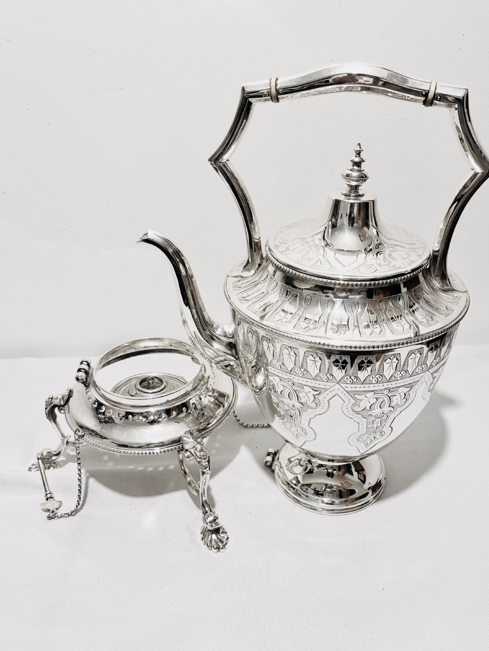 An ornate late Victorian silver-plated tea kettle with engraved geometric panels resting on a matching spirit burner stand. View II