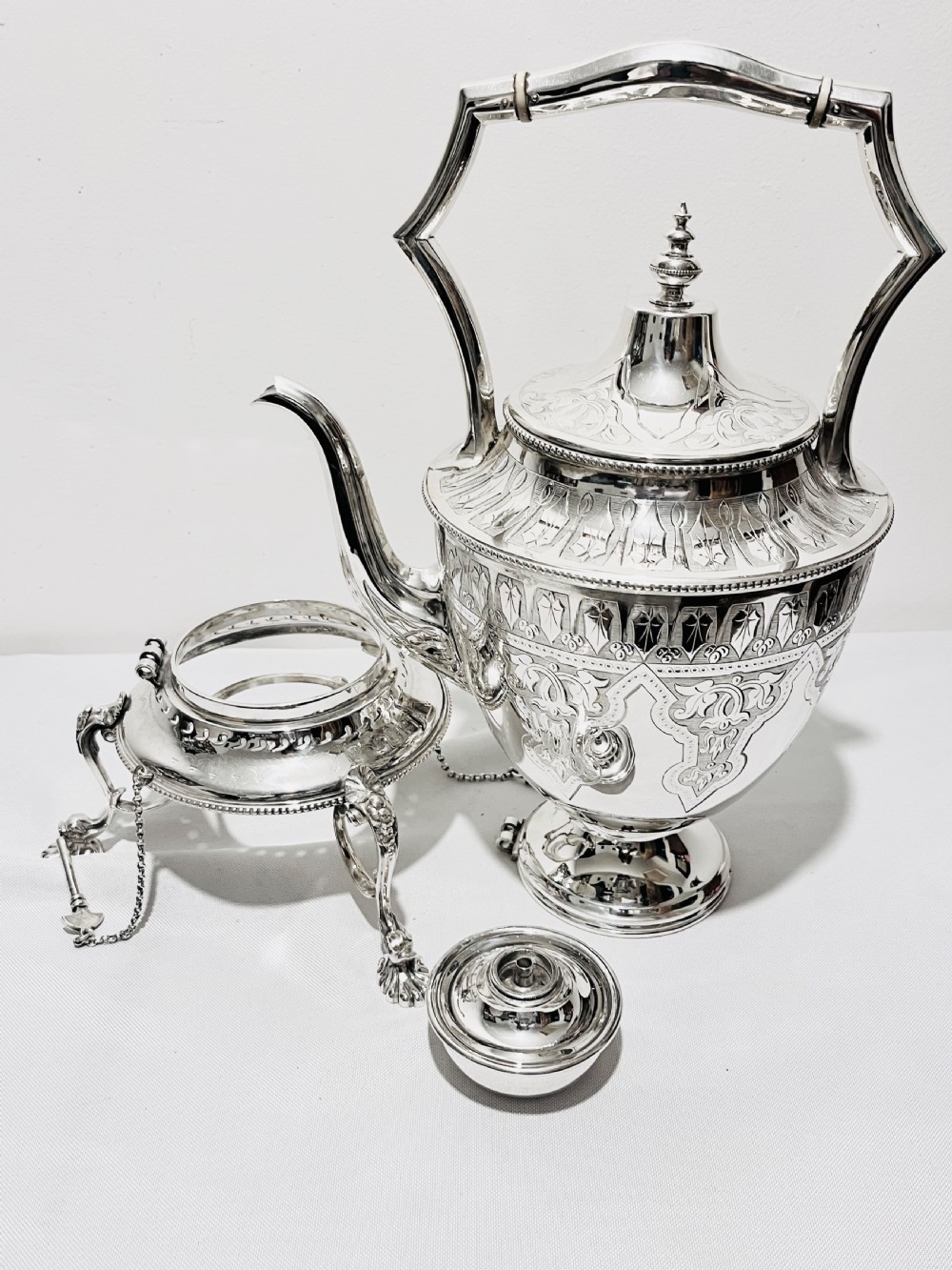 An ornate late Victorian silver-plated tea kettle with engraved geometric panels resting on a matching spirit burner stand. View III