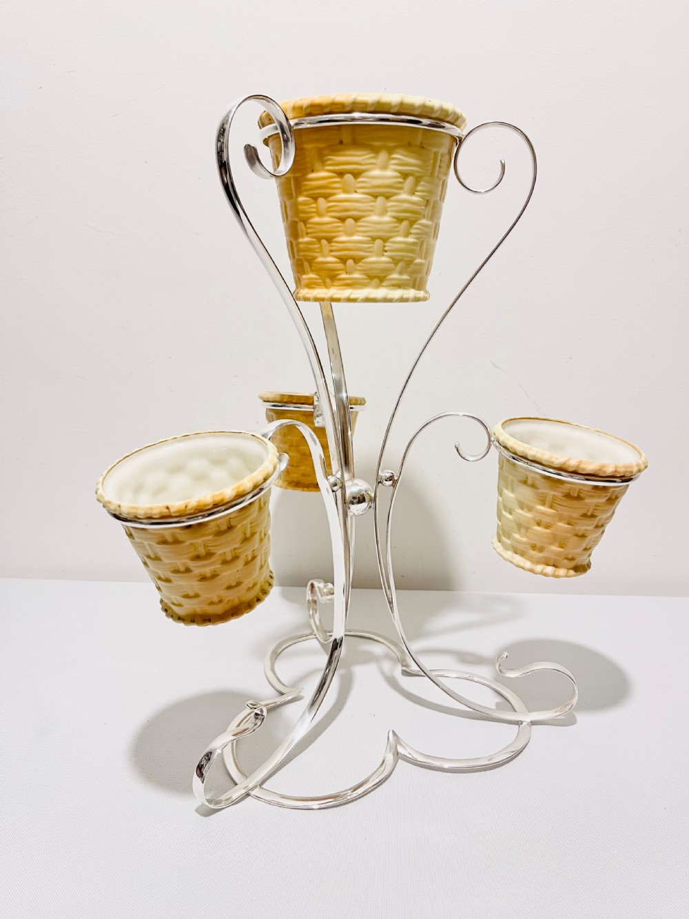 An Edwardian silver-plated tiered stand holding four cream basketweave ceramic pots on scrolling wirework supports.
