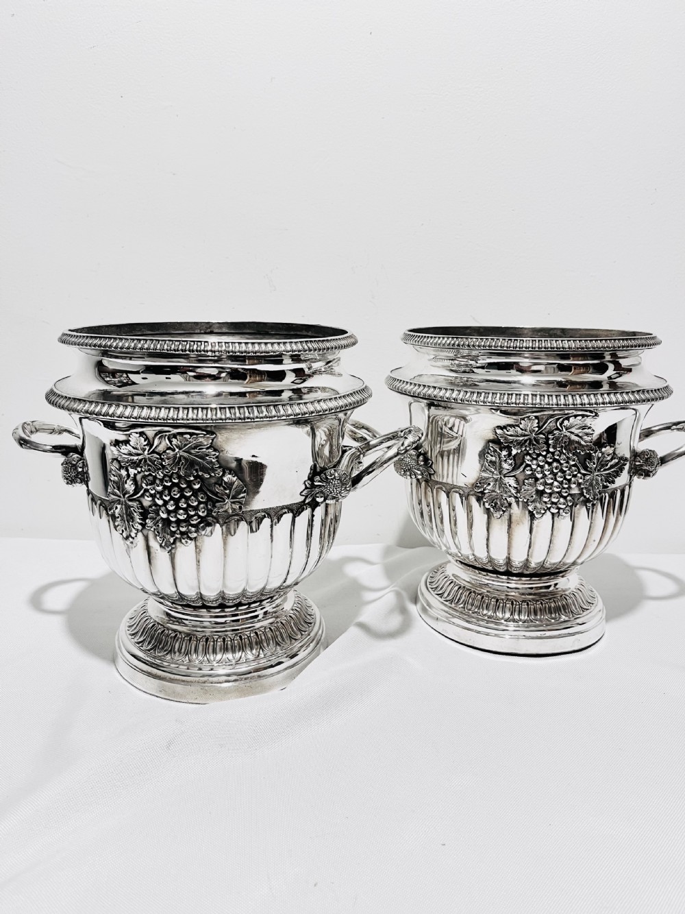 A pair of Regency silver-plated urn-shaped wine coolers featuring raised grapevine motifs, gadrooned rims, fluted bases, and twin side handles.