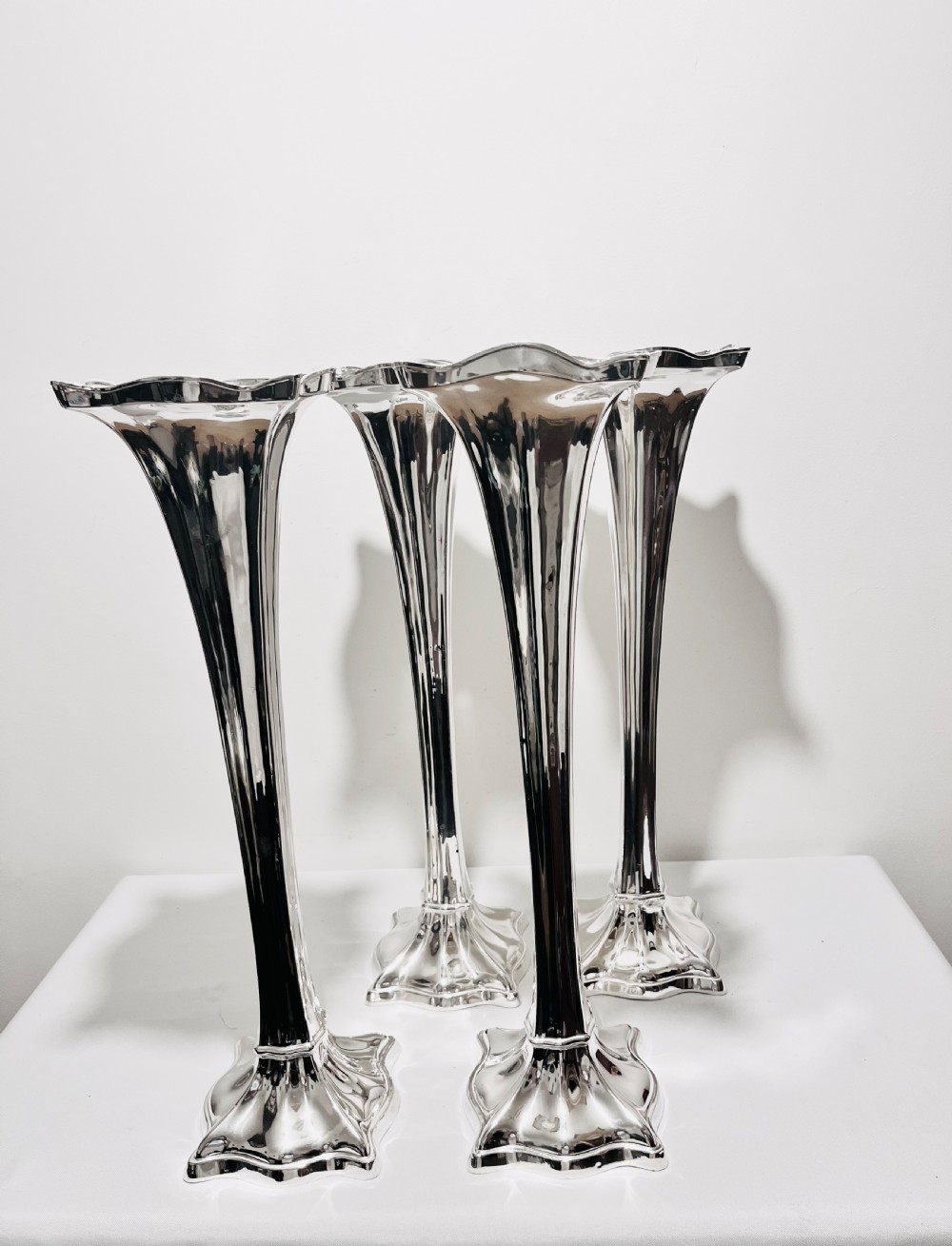 A set of four tall polished silver-plated trumpet vases featuring fluted stems, scalloped rims, and flared stepped bases.