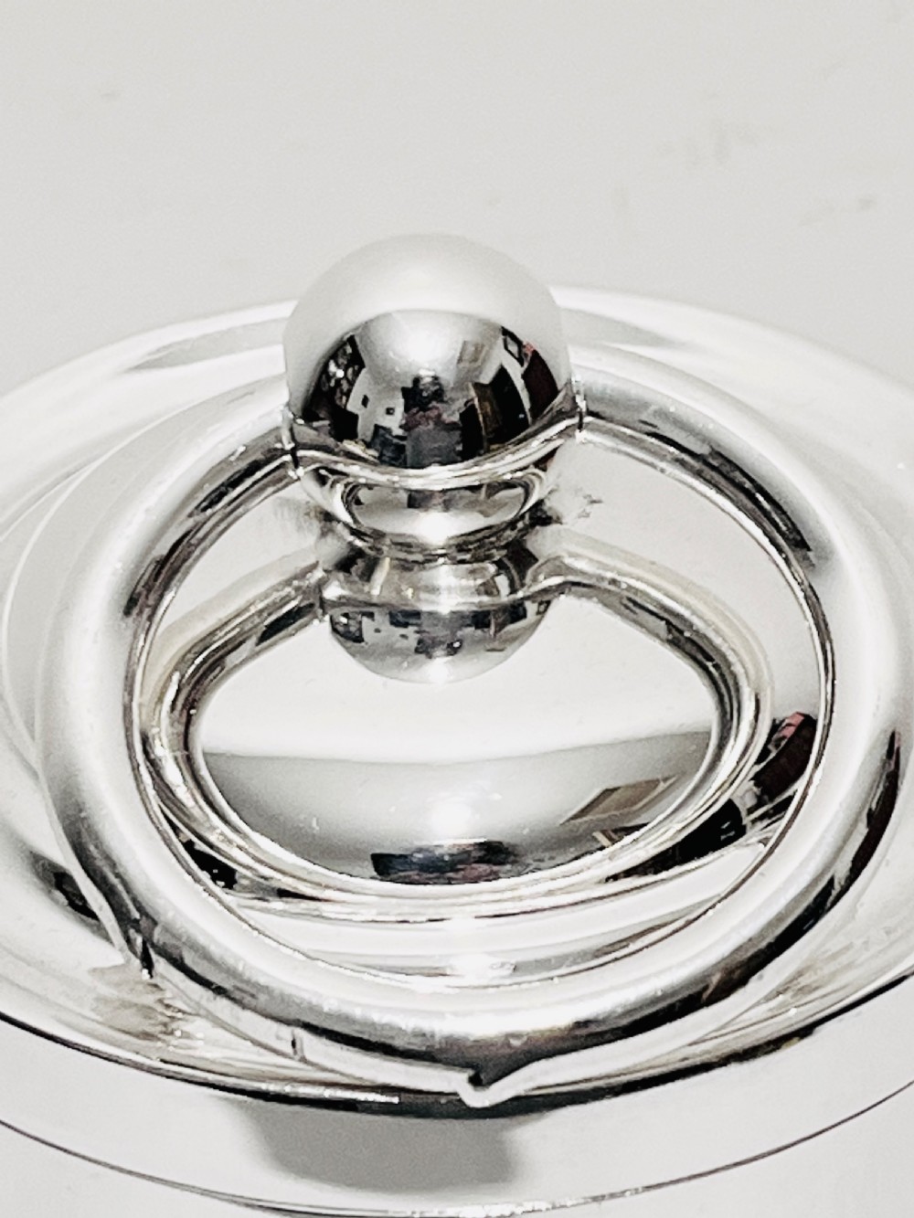 A polished silver plated cylindrical biscuit box designed in the form of a trade weight, featuring a fitted lid with a central ring handle. View II