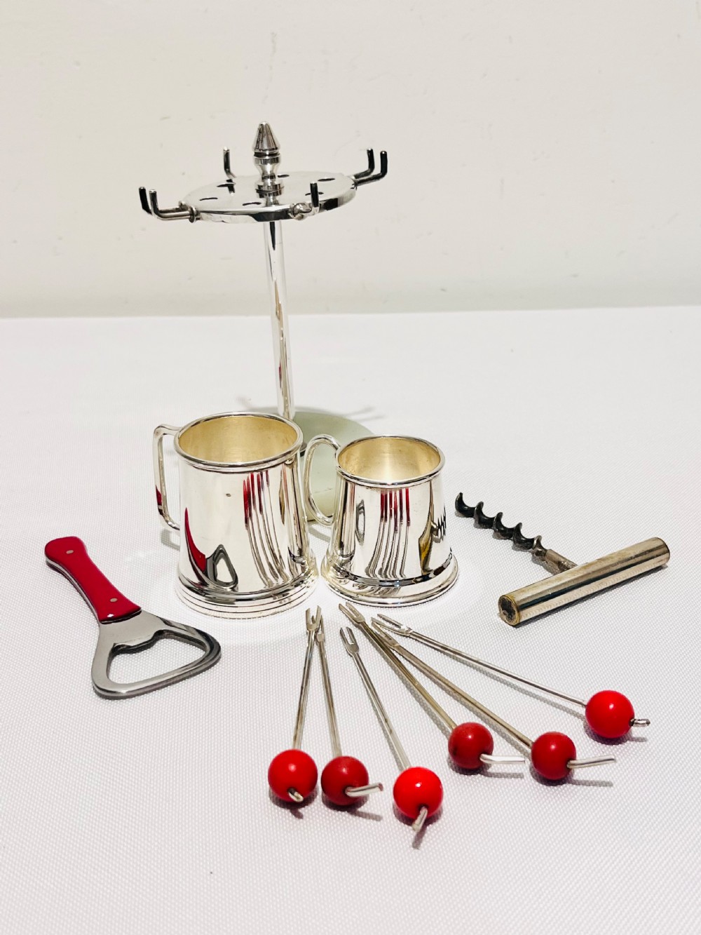 An Art Deco silver-plated bar set featuring miniature tankards, red-handled tools, and cherry cocktail picks arranged on a circular stand with a pale onyx base. View II