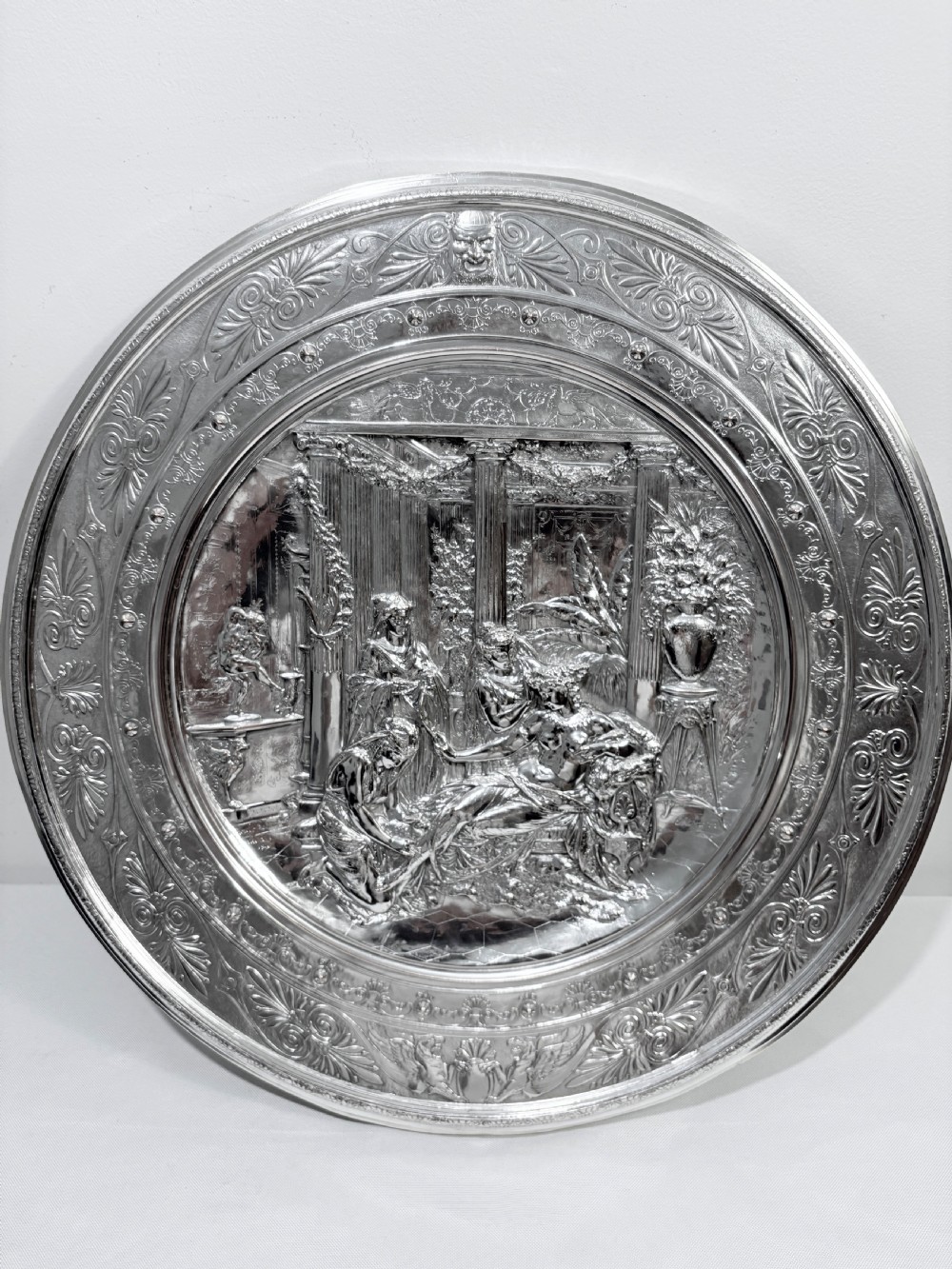 A circular silver plate electrotype charger featuring a detailed relief depiction of classical figures framed by ornate scrolled borders.