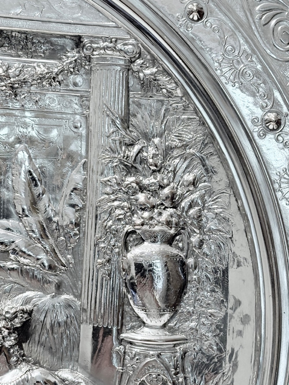 A circular silver plate electrotype charger featuring a detailed relief depiction of classical figures framed by ornate scrolled borders. View III