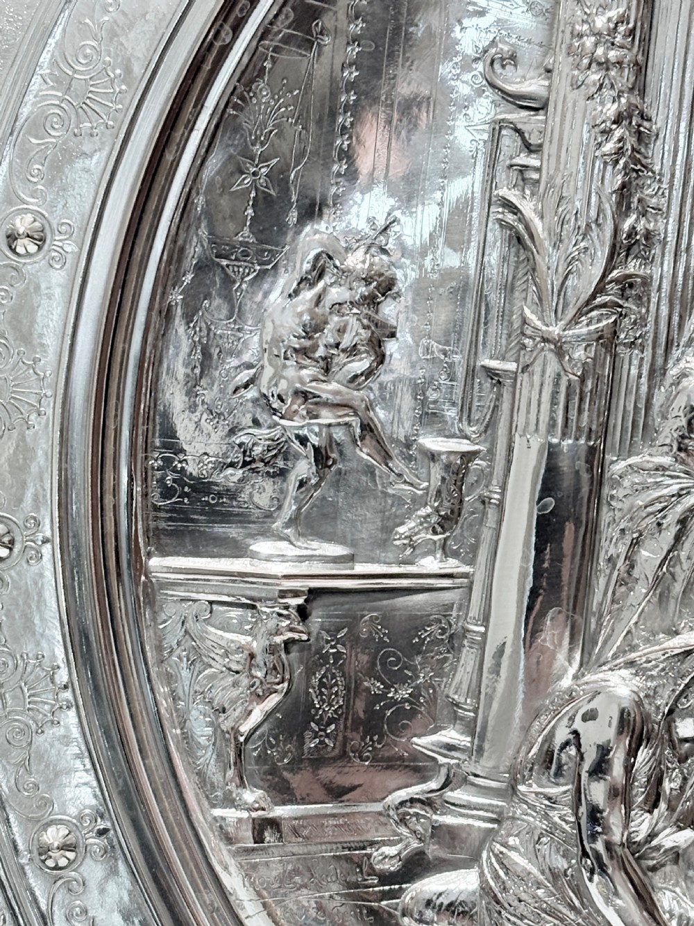 A circular silver plate electrotype charger featuring a detailed relief depiction of classical figures framed by ornate scrolled borders. View IV