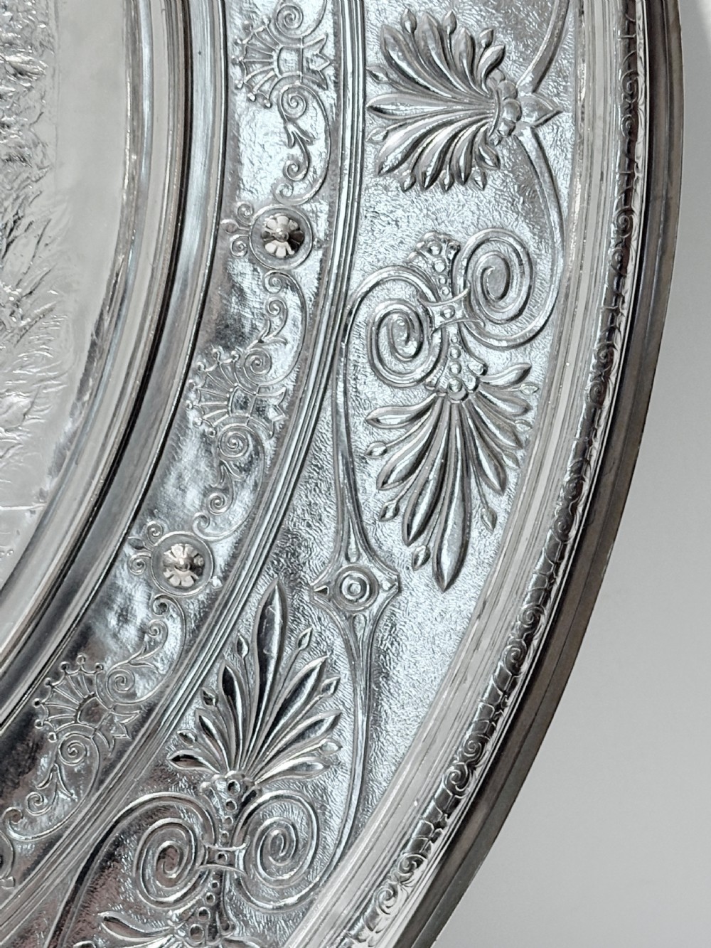 A circular silver plate electrotype charger featuring a detailed relief depiction of classical figures framed by ornate scrolled borders. View V