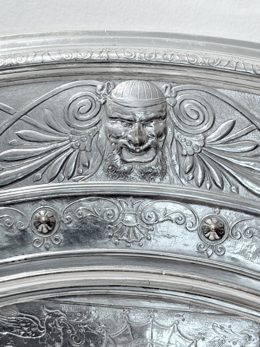 A circular silver plate electrotype charger featuring a detailed relief depiction of classical figures framed by ornate scrolled borders. View VI