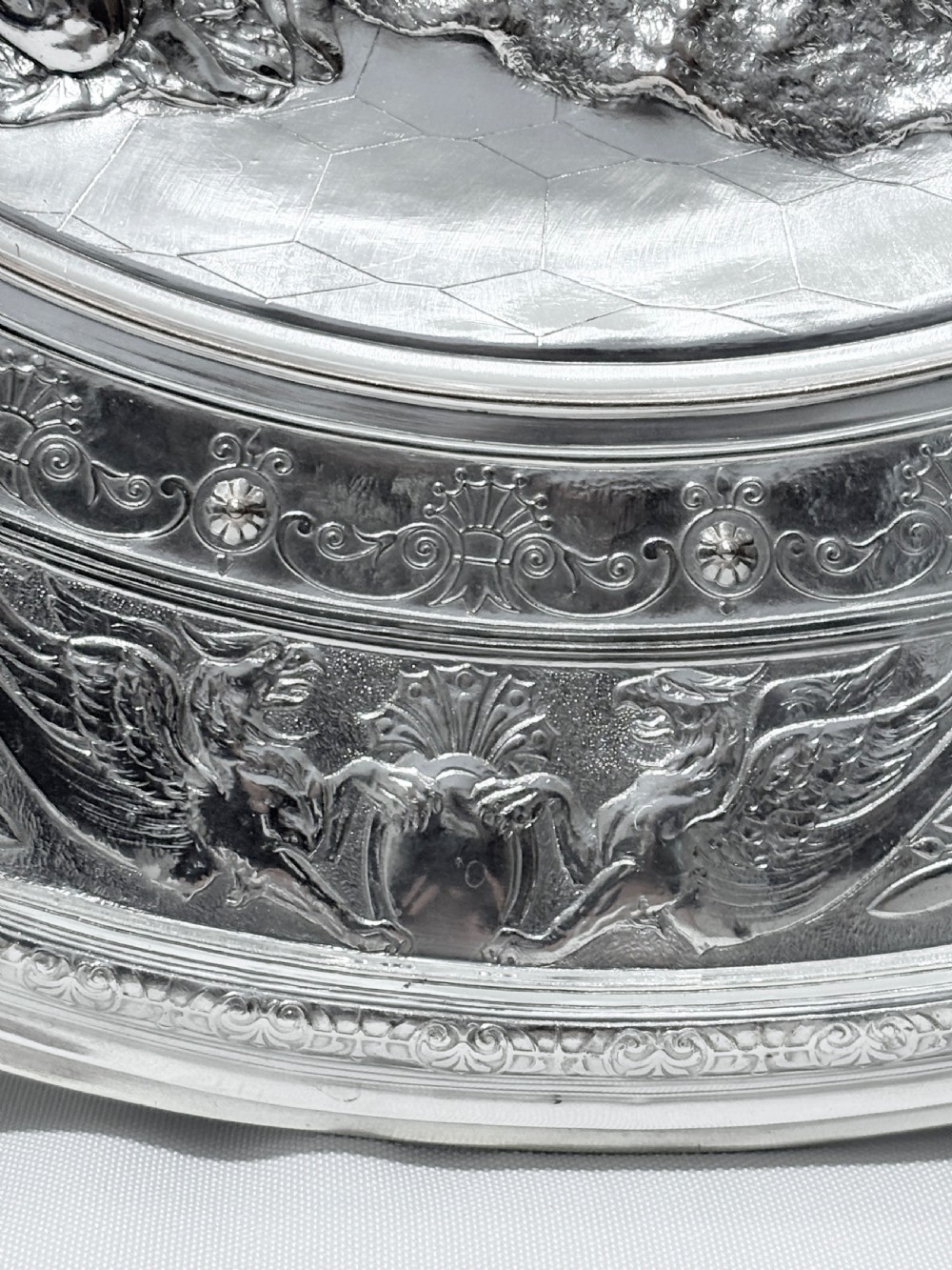 A circular silver plate electrotype charger featuring a detailed relief depiction of classical figures framed by ornate scrolled borders. View VII