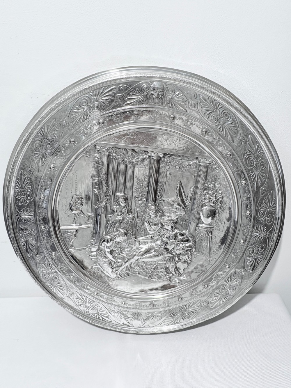 A circular silver plate electrotype charger featuring a detailed relief depiction of classical figures framed by ornate scrolled borders. View VIII