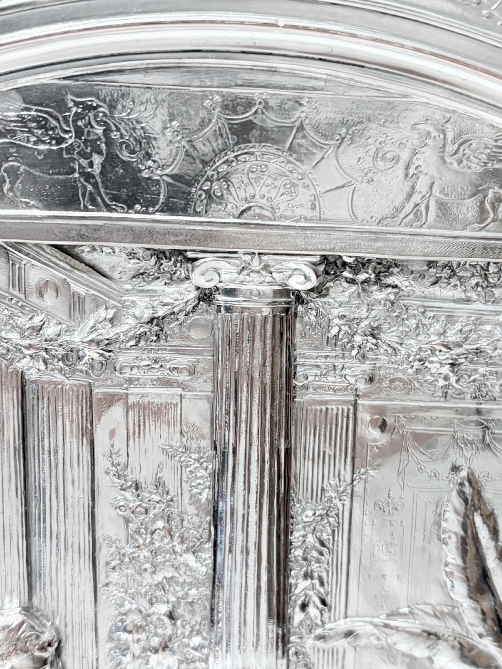 A circular silver plate electrotype charger featuring a detailed relief depiction of classical figures framed by ornate scrolled borders. View X