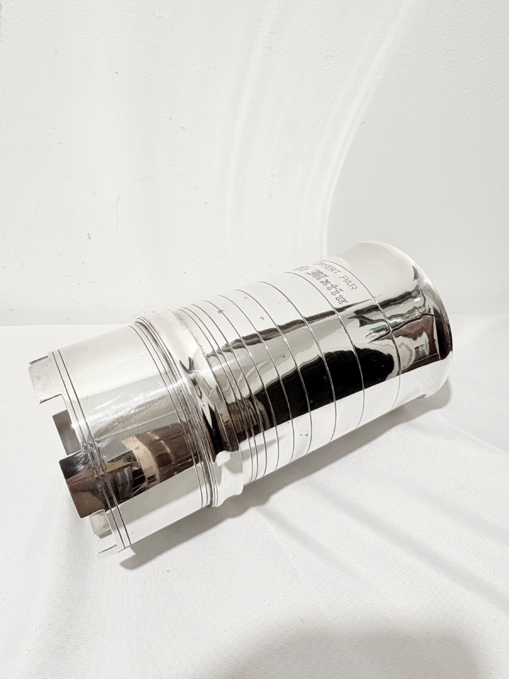 A cylindrical silver-plated wine holder featuring horizontal ring mouldings, an engraved French inscription, and a crenellated top rim. View V