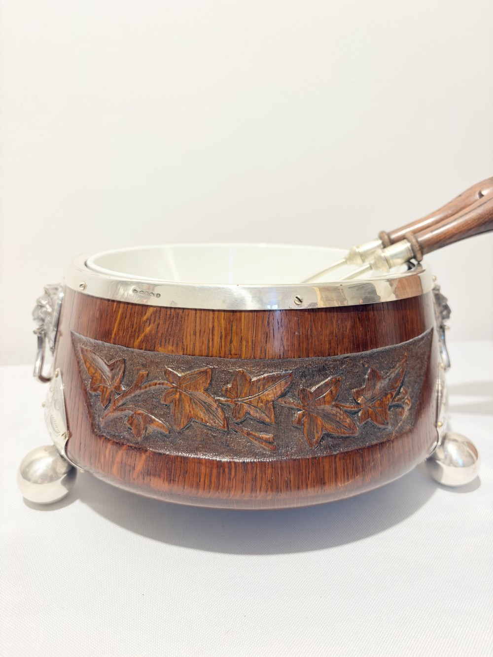 A late Victorian carved oak salad bowl with silver-plated rim, ball feet, and a white ceramic liner, featuring an ivy leaf carved band around the exterior.