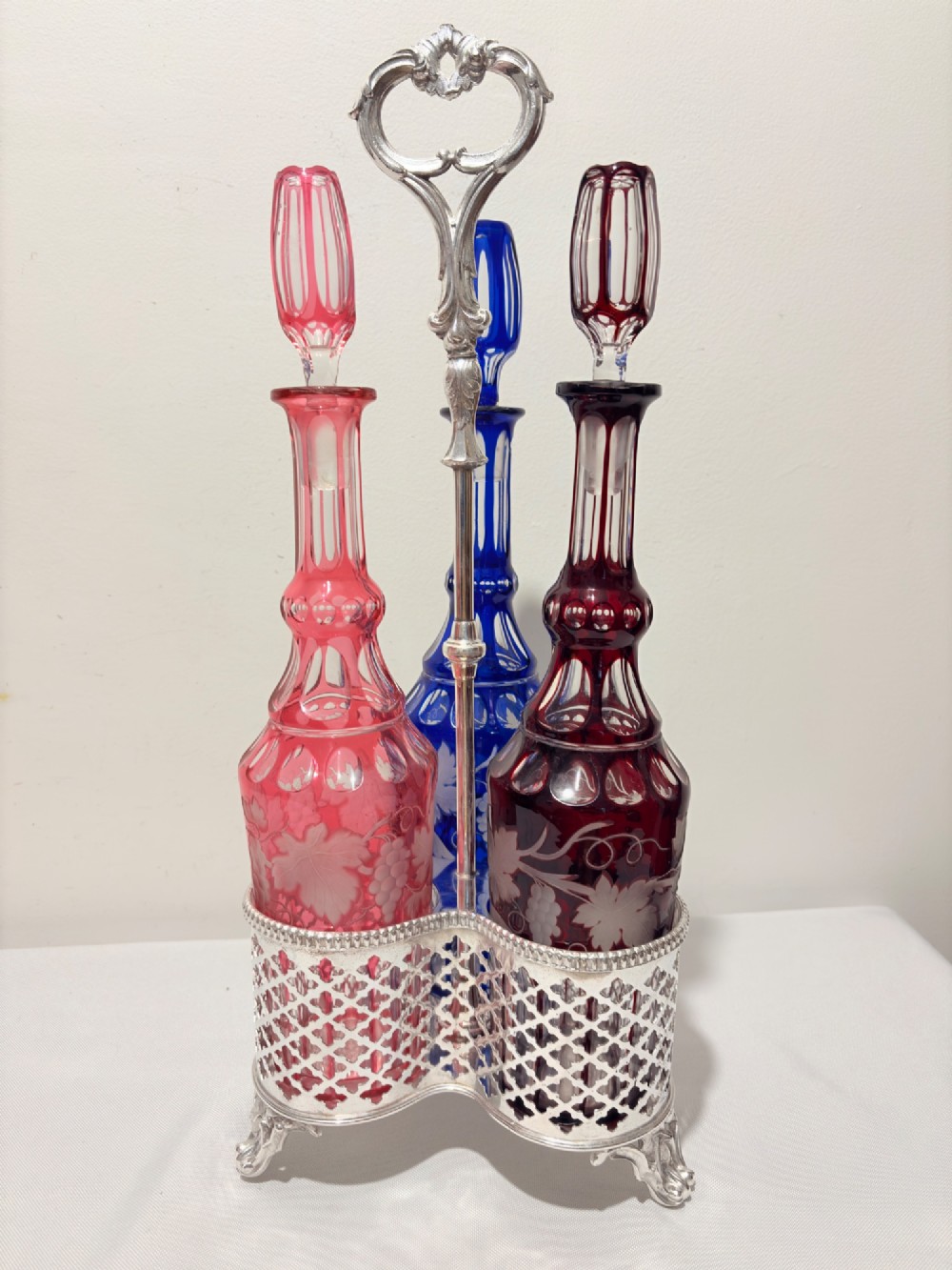 A Victorian silver-plated decanter stand holding three colourful cut-to-clear glass bottles in pink, blue, and deep red with faceted stoppers.