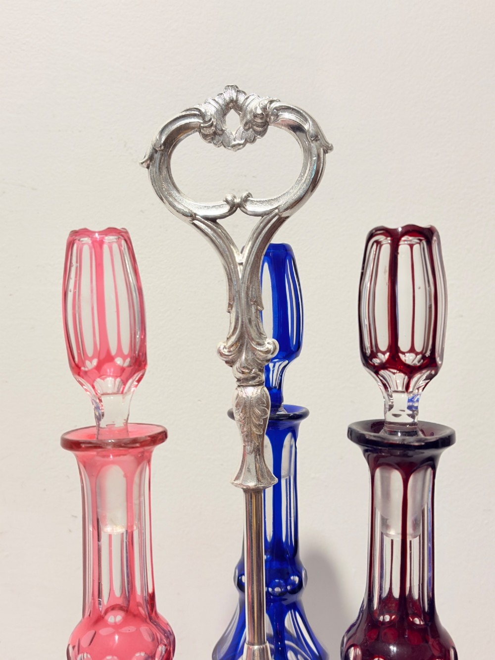 A Victorian silver-plated decanter stand holding three colourful cut-to-clear glass bottles in pink, blue, and deep red with faceted stoppers. View III