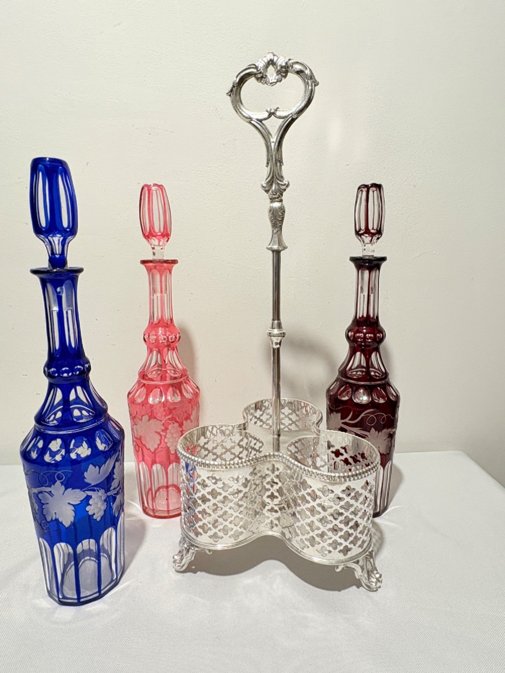 A Victorian silver-plated decanter stand holding three colourful cut-to-clear glass bottles in pink, blue, and deep red with faceted stoppers. View IV