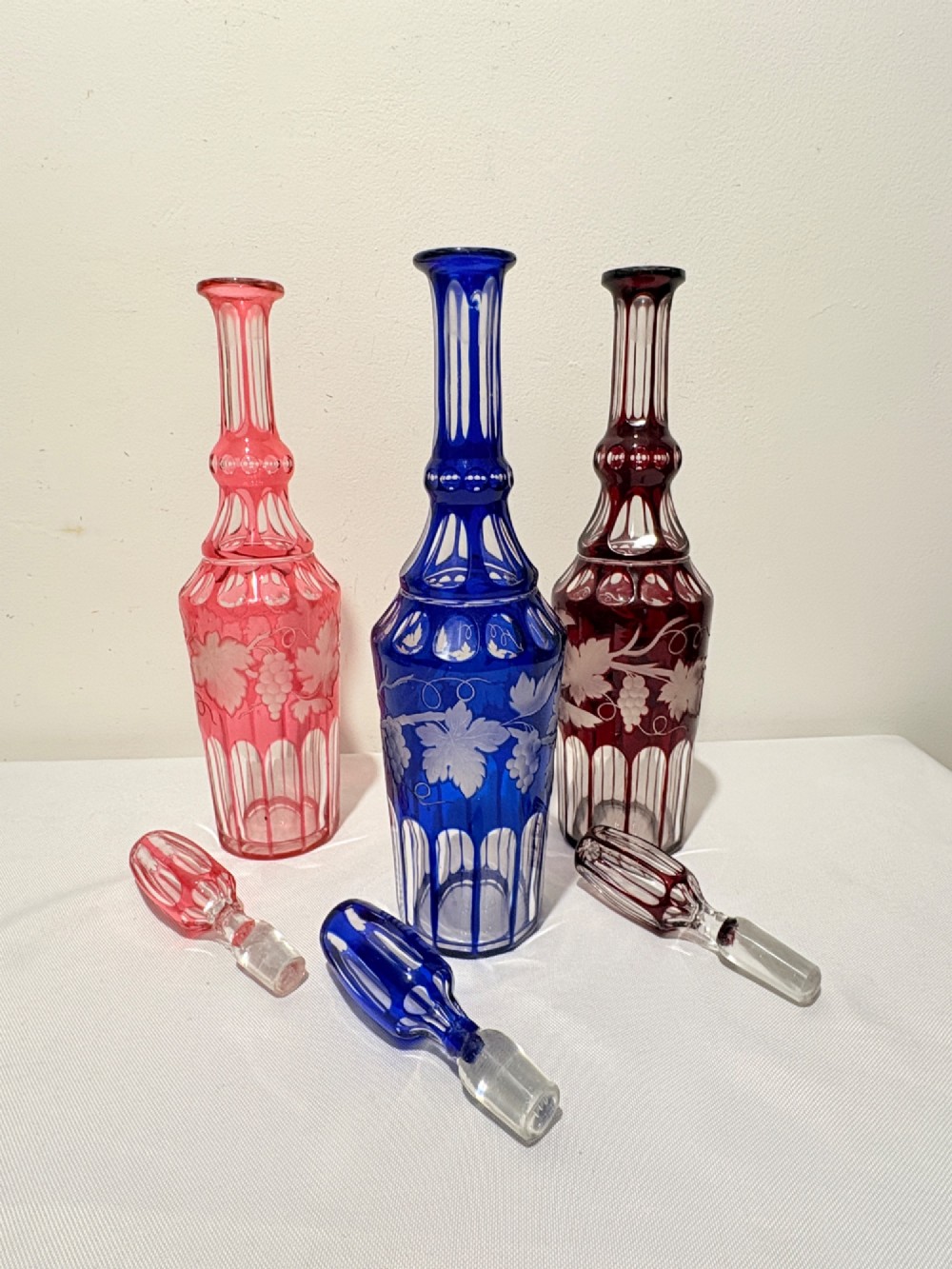 A Victorian silver-plated decanter stand holding three colourful cut-to-clear glass bottles in pink, blue, and deep red with faceted stoppers. View V