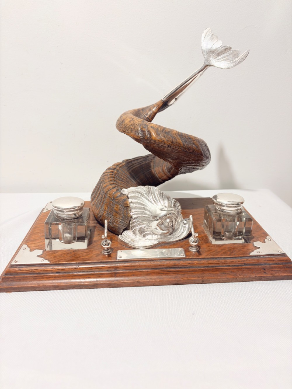 A Victorian era desk inkstand featuring a centrally mounted curled ram horn on an oak base with silver plated corner mounts and two glass inkwells.