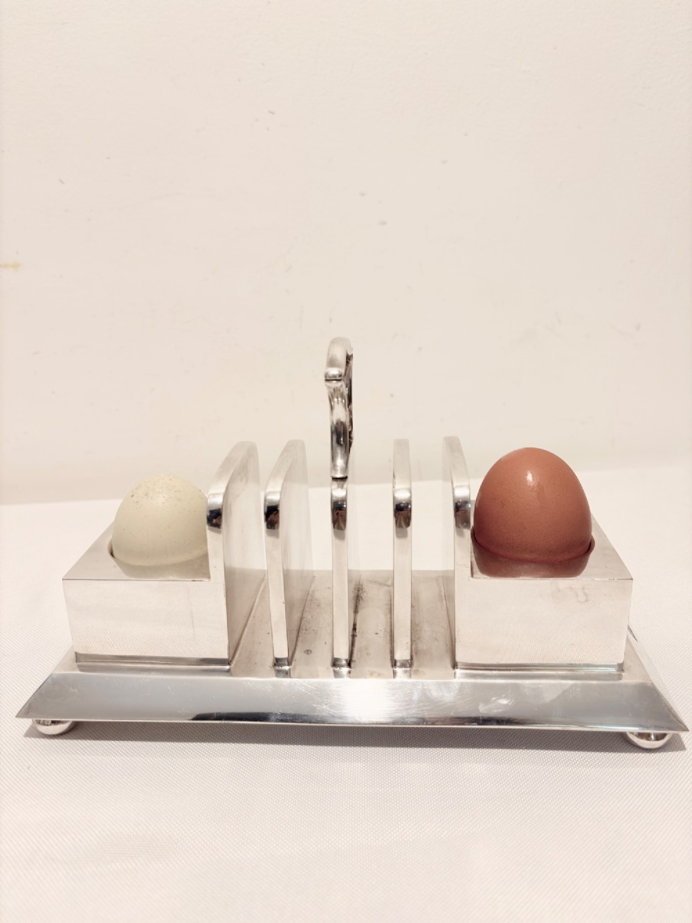 A Victorian silver-plated toast rack with radiator-style dividers flanked by two integrated egg holders on a rectangular base with ball feet. View VII