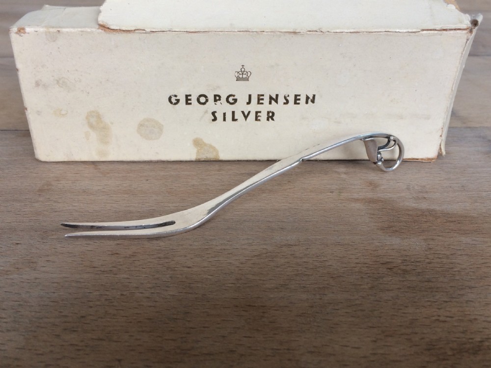 A Georg Jensen sterling silver Blossom pattern two-pronged cocktail fork resting in front of its original paper packaging box.