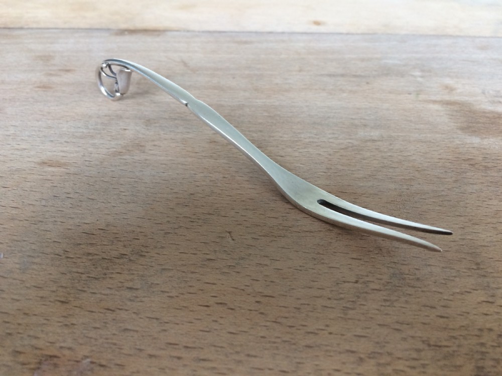 A Georg Jensen sterling silver Blossom pattern two-pronged cocktail fork resting in front of its original paper packaging box. View II