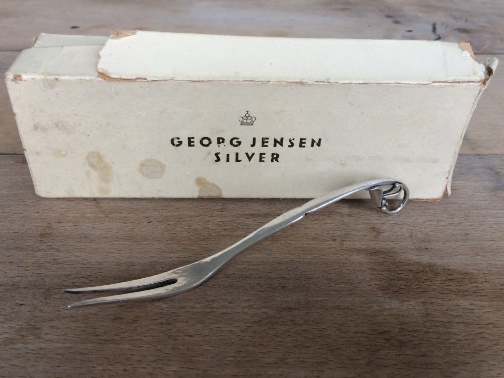 A Georg Jensen sterling silver Blossom pattern two-pronged cocktail fork resting in front of its original paper packaging box. View VI