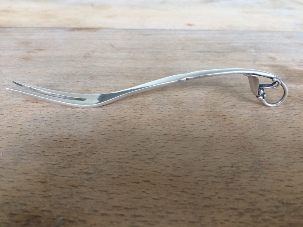 A Georg Jensen sterling silver Blossom pattern two-pronged cocktail fork resting in front of its original paper packaging box. View VIII