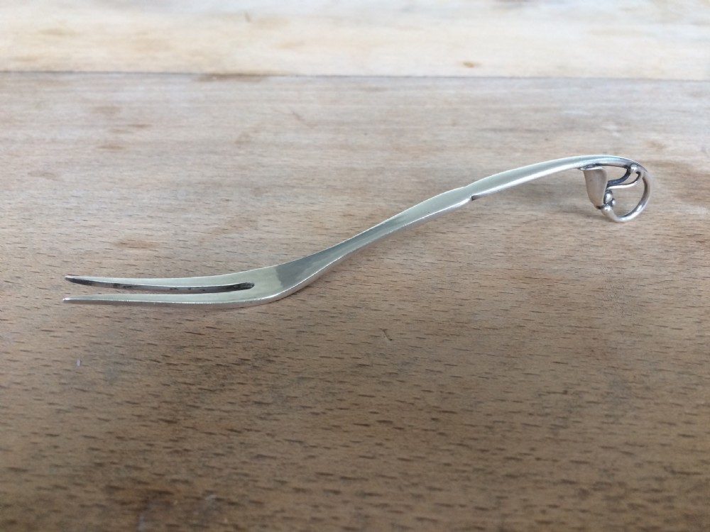 A Georg Jensen sterling silver Blossom pattern two-pronged cocktail fork resting in front of its original paper packaging box. View IX